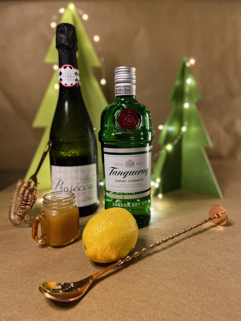 bottle of gin, bottle of fizz, lemon and shot of ginger cordial in front of a festive background