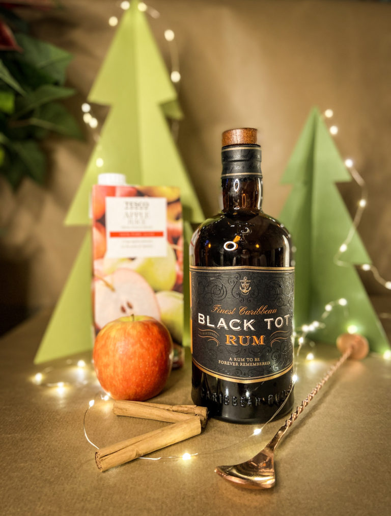 bottle of rum, apple juice, cinnamon sticks, apple and a spoon in front of a festive background