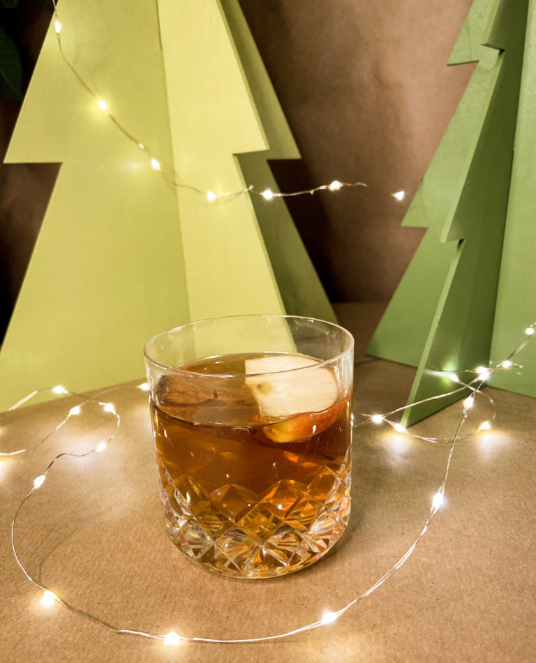hot spiced rum and apple cocktail in a rocks glass garnished with a slice of apple in front of a festive background